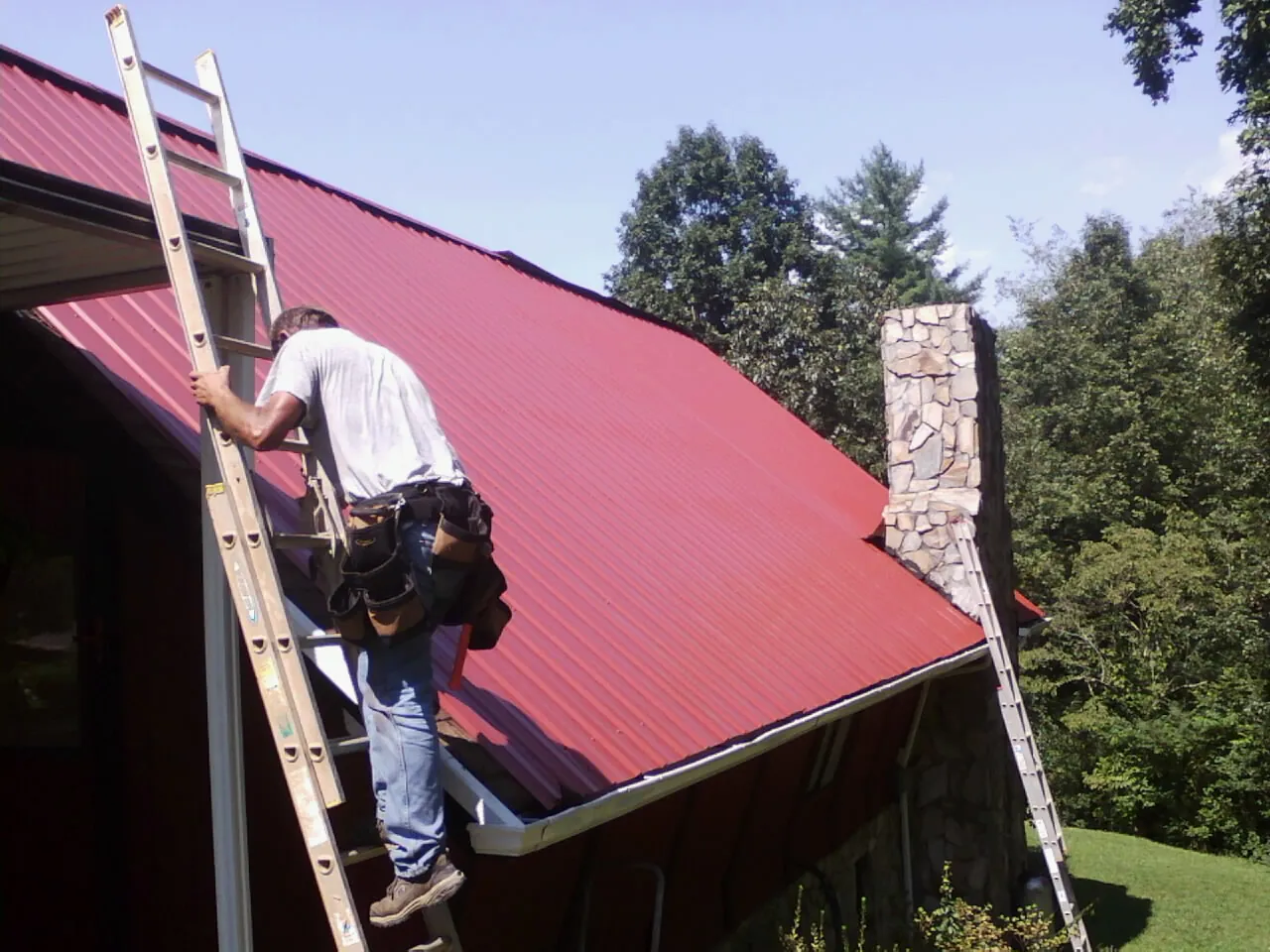 Roofer on a ladder beside a red metal roof and stone chimney — Soffit & Fascia Repair by Roofing Brothers in Quantico